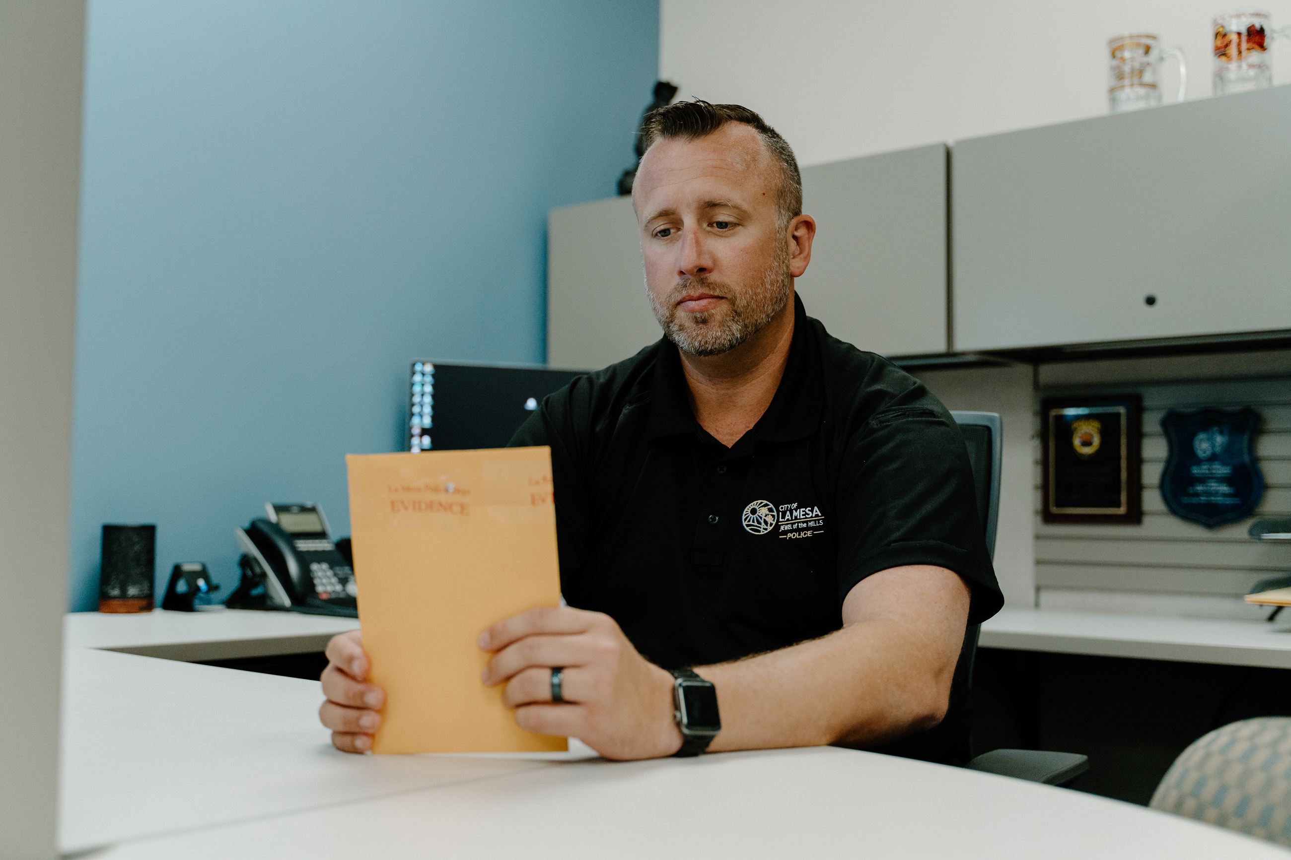 A police detective sitting at a desk holding an envelope marked as evidence.